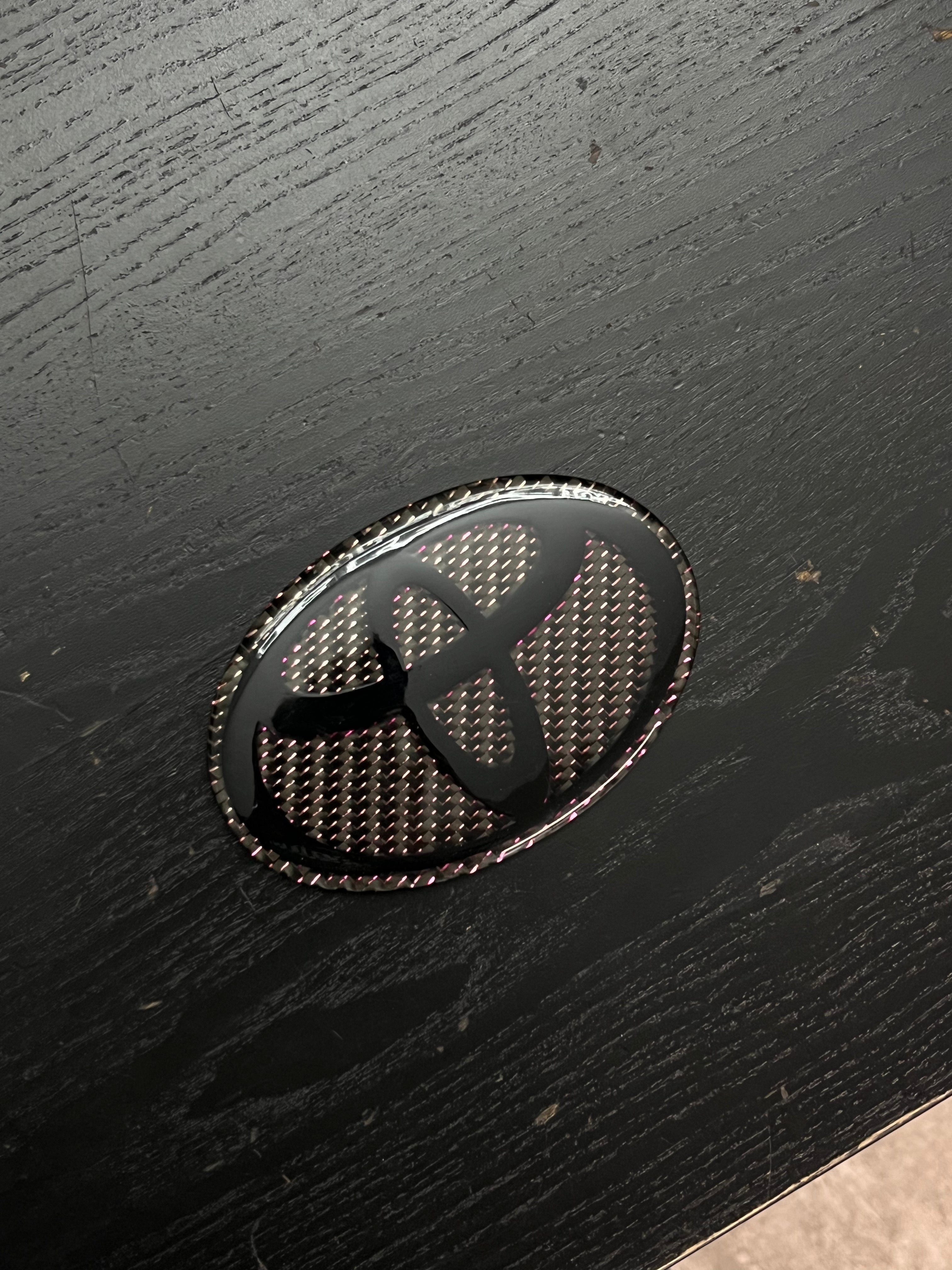 Custom Carbon Fiber Toyota Emblem - Gloss Black Logo with Pink Iridescent Weave.