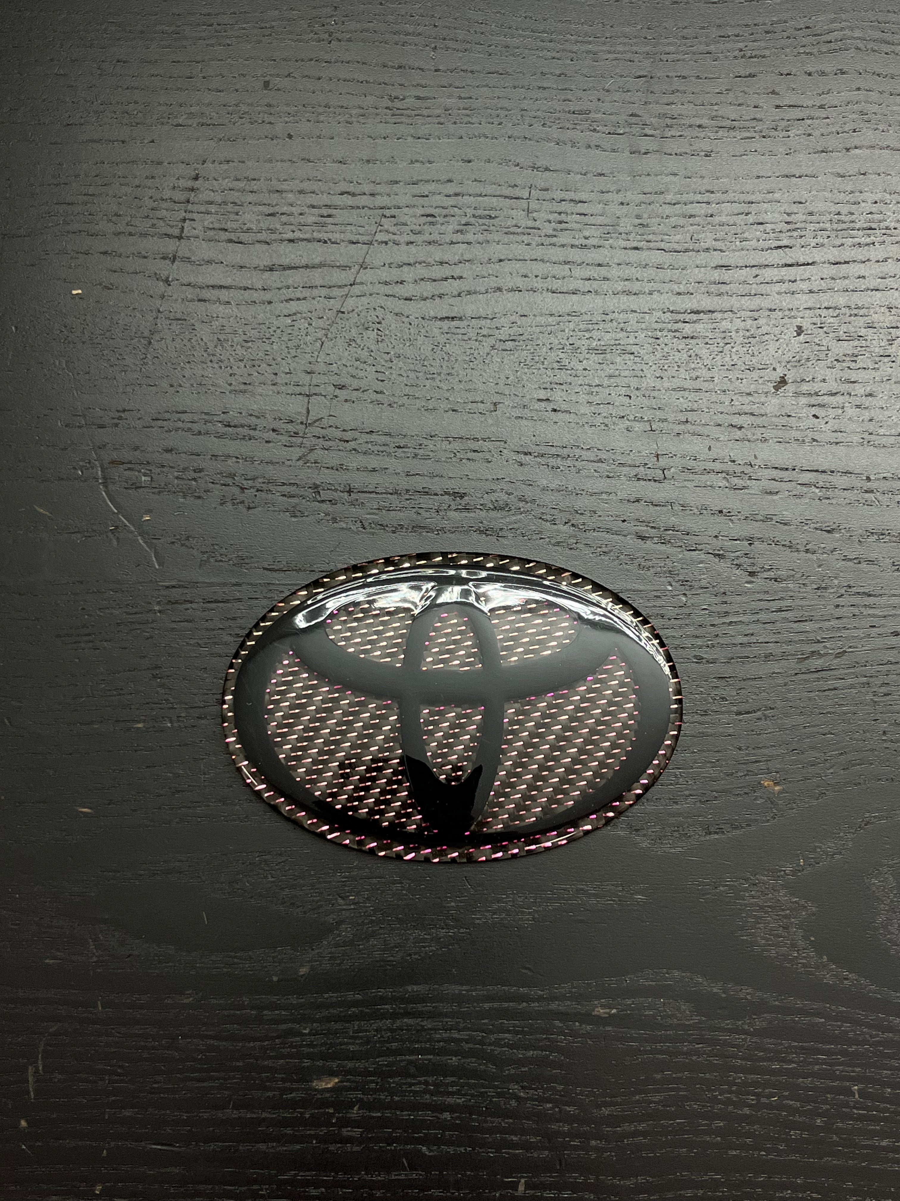 Custom Carbon Fiber Toyota Emblem - Gloss Black Logo with Pink Iridescent Weave.